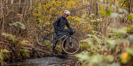Mann durchkreuzt einen Bach mit Gravel Bike und Taschen dran